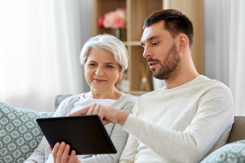 a young man sits on the couch with an older woman and points to a tablet while discussing how she can prepare for dental implant surgery in Oshkosh 